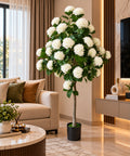 Artificial white hydrangea tree with abundant white flower clusters and lush green leaves in a black pot, displayed in a living room.